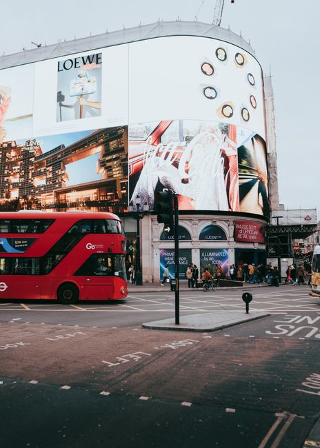 A large curved digital advertising display on a building in Soho, London, featuring multiple screens with various images and branding, including the word 'LOEWE' at the top. In front of the building, 