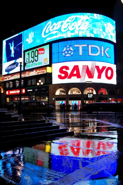 A large curved digital advertising display on a building in Soho, London, featuring multiple screens with various images and branding, including the word 'LOEWE' at the top. In front of the building, 